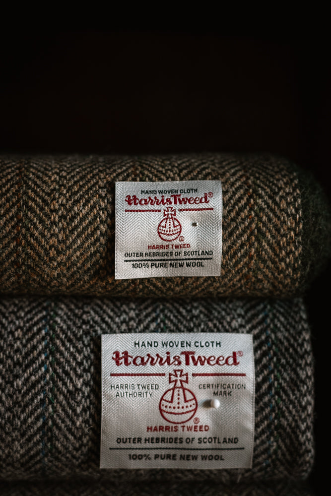 Two rolls of House Tweed 95054 by Campbell’s of Beauly, each featuring brown and green patterned fabric with a white label bearing the Harris Tweed Authority orb logo and certifying it is handwoven in Scotland from 100% pure new wool. These distinguished jacketing cloths boast a classic herringbone design, perfect for timeless elegance.
