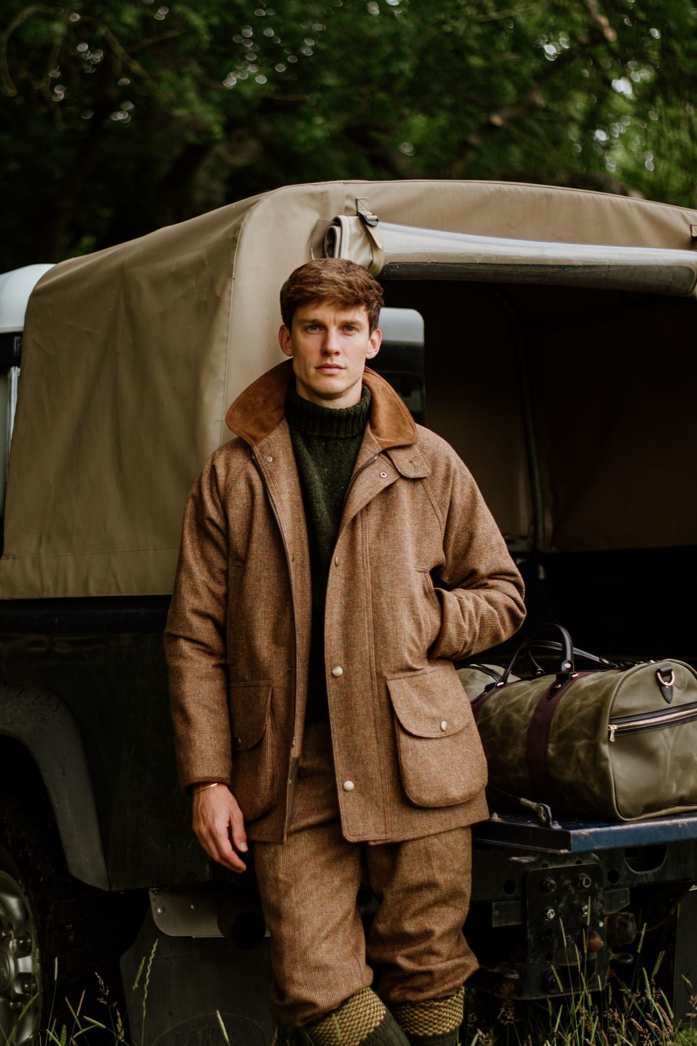A man in brown outdoor gear stands by a vehicle with a tan canvas top, one hand in his pocket. The Campbells of Beauly Wax Holdall rests on the tailgate—your ideal travel companion—against a backdrop of lush greenery.
