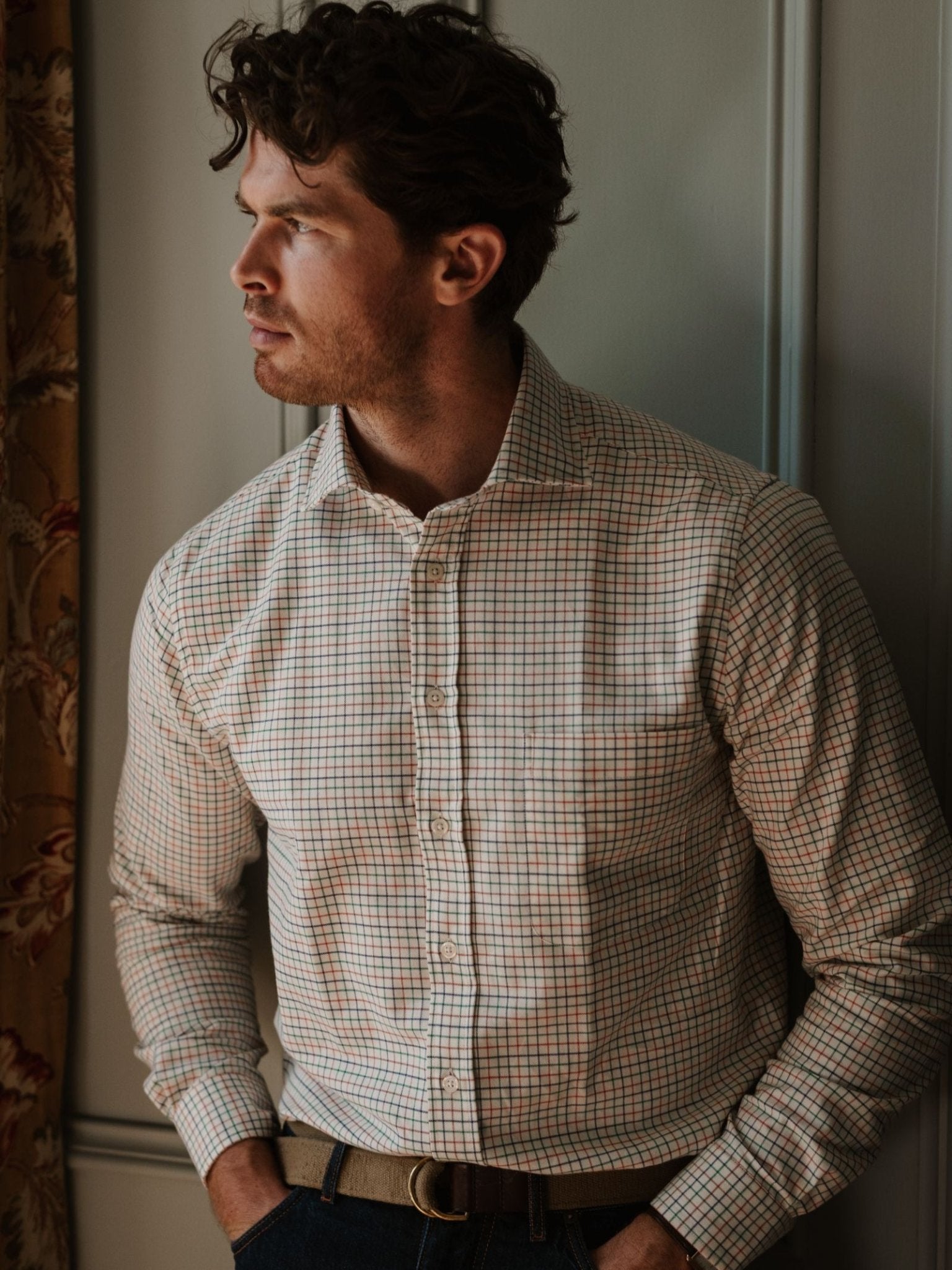 A man stands indoors, looking thoughtfully to the side, dressed in a light-colored Campbell's of Beauly Windowpane Shirt with dark jeans and a tan belt, his face softly lit from the left.