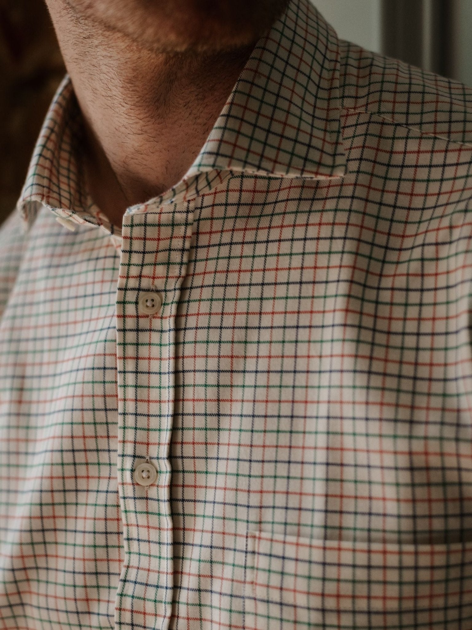 A close-up of a person wearing the Campbell's of Beauly Windowpane Shirt, a beige brushed cotton shirt with a red, green, and blue checkered pattern. The shirt is buttoned up, showing the wearer’s neck and part of their chin.
