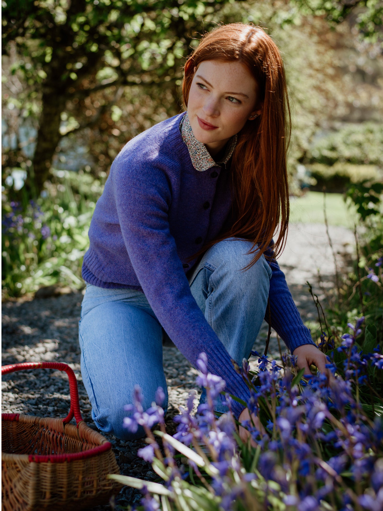 A young woman with long red hair, wearing the Campbells of Beauly Crew Cardigan and light blue jeans, kneels on a gravel path among blooming purple flowers. She reaches for the flowers, a basket beside her amidst sunlight and greenery.