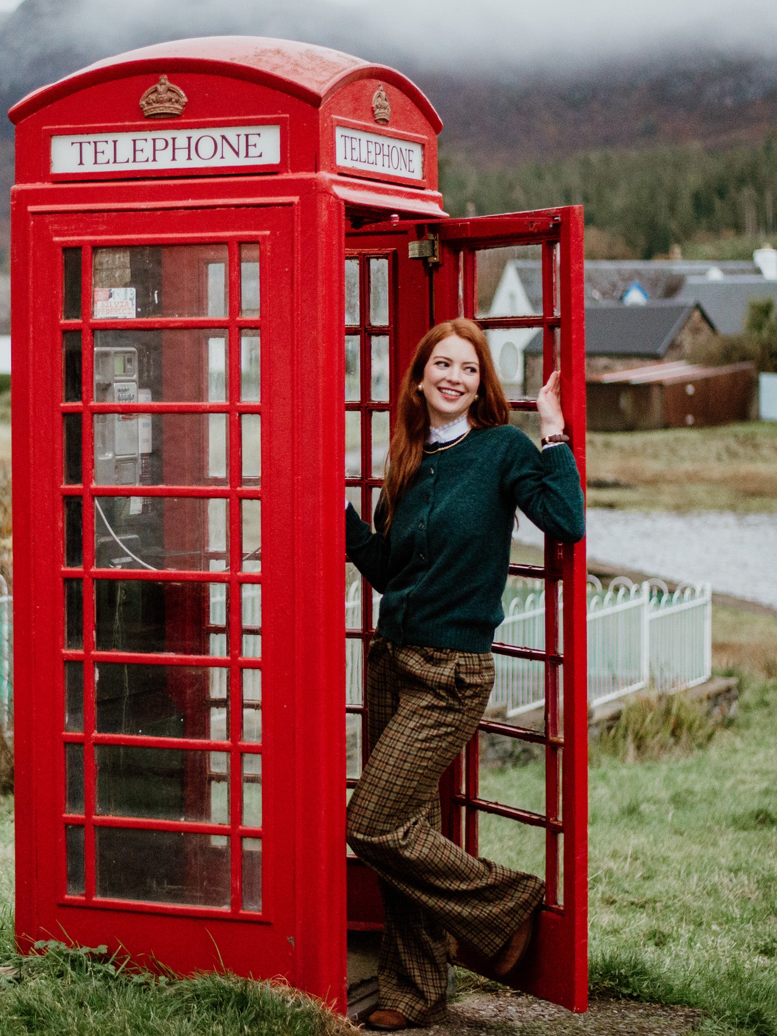 A woman with long red hair, wearing a dark green Campbells of Beauly Crew Cardigan and plaid pants, smiles as she leans against the open door of a classic red British phone booth in a rural landscape with grass, houses, and mountains behind her.